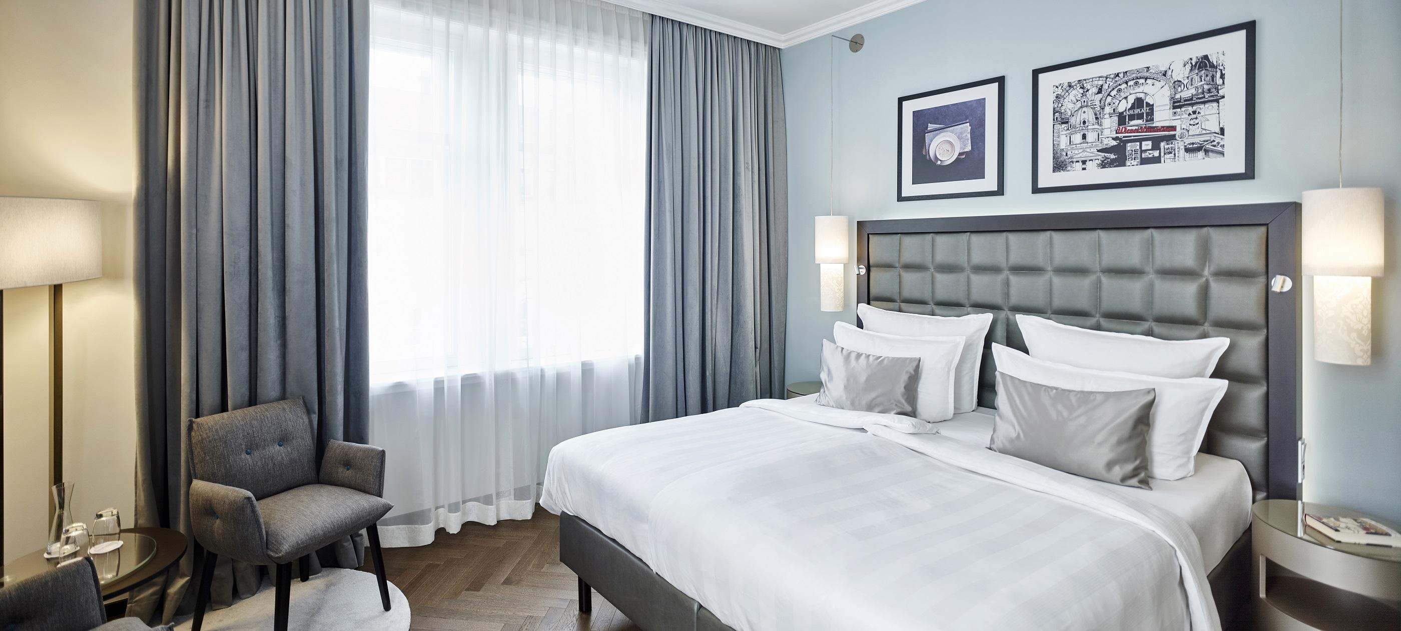 Steigenberger Hotel Herrenhof Вена Экстерьер фото The photo shows a modern hotel room featuring a neatly made double bed with white bedding and decorative gray pillows. Behind the bed, there is a stylish, tufted headboard. To one side, there's a comfortable armchair next to a small round table. The 