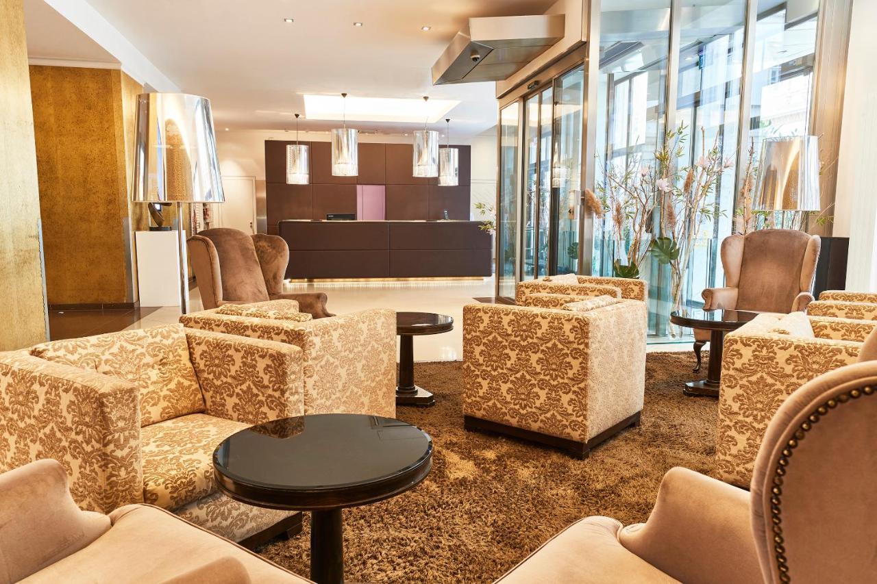 Steigenberger Hotel Herrenhof Вена Экстерьер фото The photo shows a hotel lobby or waiting area designed with a modern and elegant style. There are several plush, patterned armchairs arranged around small round tables. The flooring is covered with a large, soft rug. In the background, there is a rec